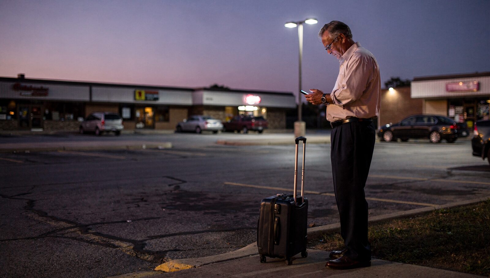 A middle-aged Ohio business traveler standing next to his rolling suitcase on the curb of a strip mall parking lot at 5:30 AM before dawn.